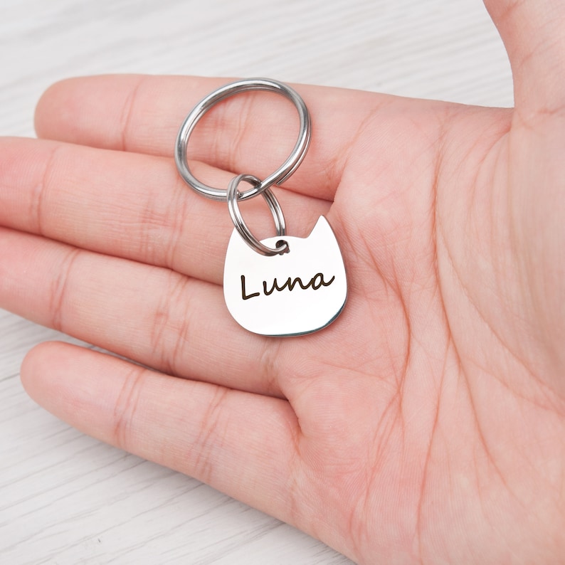 Puede incluir: Una placa de identificaci&oacute;n para mascotas de metal plateado con forma de cabeza de gato y el nombre Luna grabado en una fuente cursiva oscura. La placa est&aacute; unida a una anilla met&aacute;lica circular, formando un llavero o accesorio de identificaci&oacute;n.