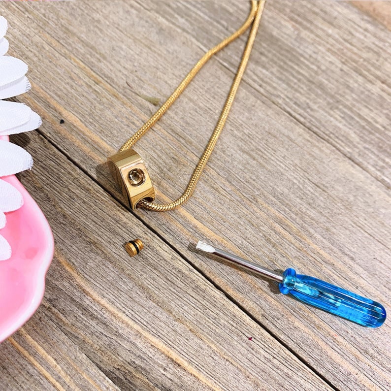 Puede incluir: Un collar de cadena dorada con un peque&ntilde;o colgante de forma cuadrada. El colgante tiene una parte superior con tornillo y se incluye un peque&ntilde;o destornillador. El colgante es probablemente un collar de cremaci&oacute;n.