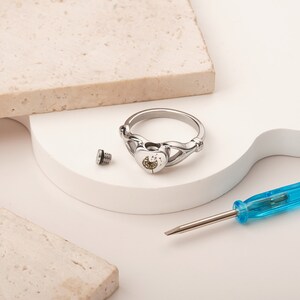May include: A silver ring with a heart-shaped design featuring a dandelion inside. The ring is open and a small screw is lying next to it. A blue screwdriver is also in the image.