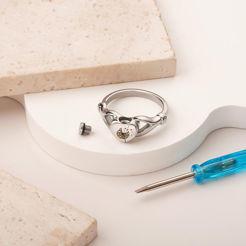 May include: A silver ring with a heart-shaped design featuring a dandelion inside. The ring is open and a small screw is lying next to it. A blue screwdriver is also in the image.
