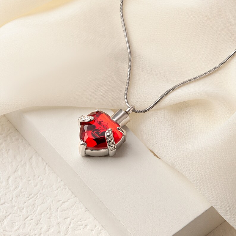 May include: A silver chain necklace with a red heart-shaped pendant. The pendant has a silver trim and is engraved with the words "Always in my heart".
