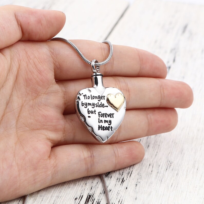 May include: Silver heart-shaped cremation necklace with a gold chain. The pendant has an inscription that reads "No longer by my side... but forever in my heart".