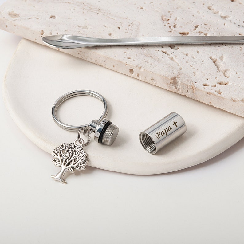 May include: Silver keychain with a small, engraved cylinder that reads "Papa +" and a tree charm. The cylinder is open and the keychain is on a white surface.