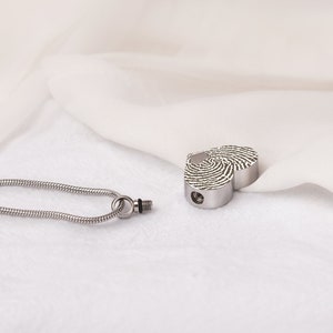 Puede incluir: Un colgante de cremaci&oacute;n de plata en forma de coraz&oacute;n con un dise&ntilde;o de huella digital. El colgante est&aacute; unido a una cadena de plata.