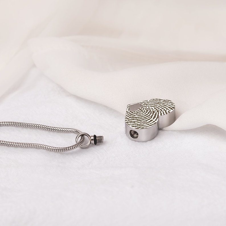 Puede incluir: Un colgante de cremaci&oacute;n de plata en forma de coraz&oacute;n con un dise&ntilde;o de huella digital. El colgante est&aacute; unido a una cadena de plata.