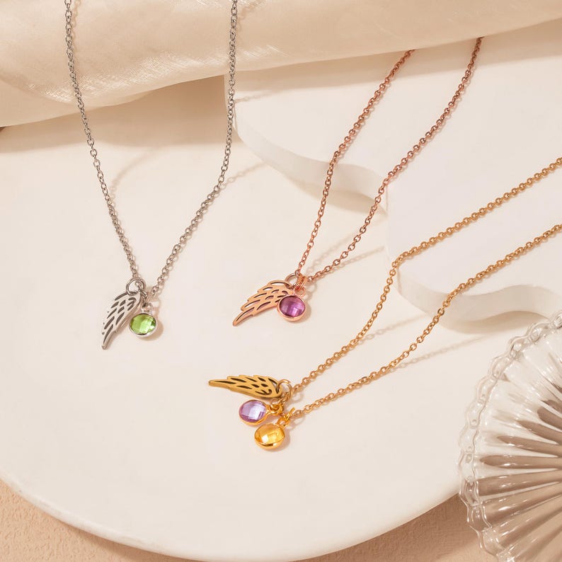 May include: Three necklaces with wing pendants and gemstone accents. One necklace is silver with a green gemstone, another is rose gold with a pink gemstone, and the third is gold with purple and yellow gemstones.
