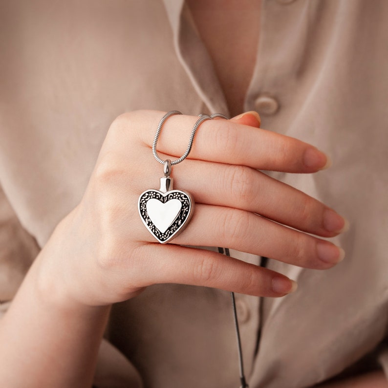 May include: A silver heart-shaped pendant necklace with a delicate chain. The pendant has a detailed, ornate design around the edge and a smooth, white center.