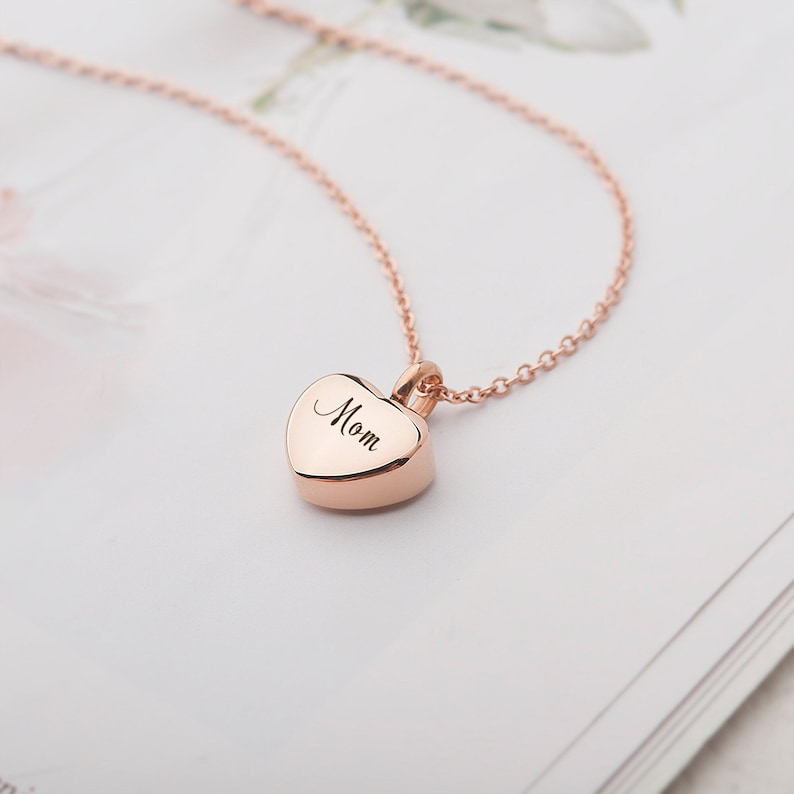 May include: A rose gold heart-shaped pendant necklace with the word "Mom" engraved on it.