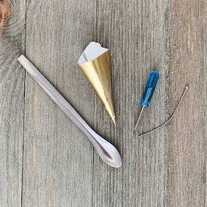 May include: A gold and white paper cone, a silver metal spoon, a blue screwdriver, and a silver wire on a wood background.