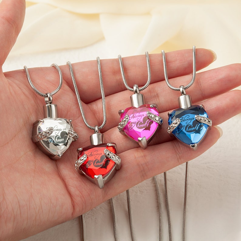 May include: Four silver heart-shaped cremation necklaces with different colored crystals. Each pendant has the inscription "Always in my heart".