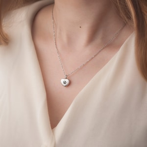 May include: A silver necklace with a heart-shaped pendant featuring a paw print cutout. The necklace is worn by a person with a white shirt.