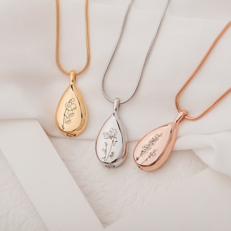 May include: Three teardrop-shaped cremation jewelry pendants on chains. The pendants are engraved with different floral designs. The chains are gold, silver, and rose gold.