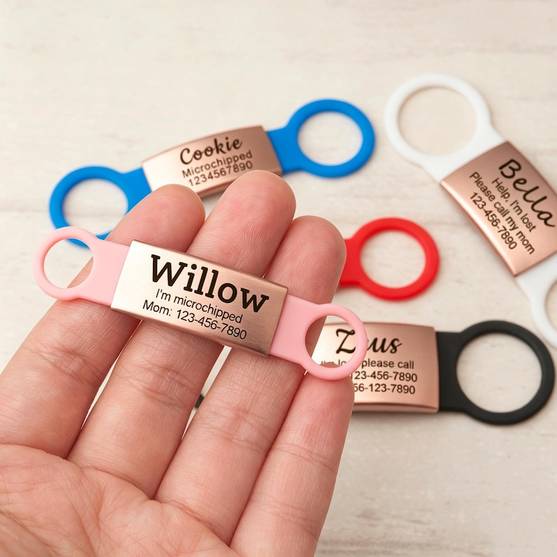 May include: A rectangular rose gold metal pet ID tag with a pink silicone band featuring the name Willow, the text I'm microchipped, and the phone number 123-456-7890. Additional tags in blue, white, red, and black are visible in the background.