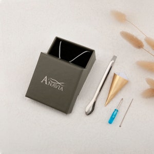 May include: A silver necklace in a dark green box with the word "Anavia" printed on the box. The box is on a white surface with a gold and white paper cone, a silver tool, a blue screwdriver, and a silver needle.