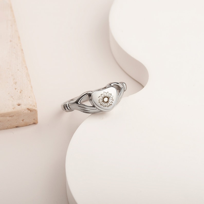 May include: Silver ring with a heart-shaped design. The heart features a small flower design inside. The ring is designed to hold a small amount of ashes or other mementos.