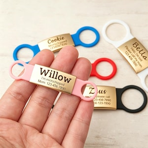 May include: A collection of rectangular gold-toned metal pet ID tags with silicone loops in pink, blue, white, red, and black. Each tag is engraved with a pet name, such as Willow, Cookie, Bella, or Zeus, along with contact information.