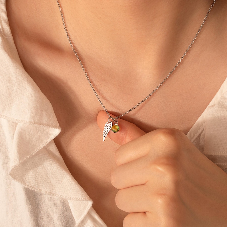 May include: Silver necklace with a delicate chain. The pendant features a small wing charm and a round, yellow-green gemstone. The necklace is worn against a white shirt.