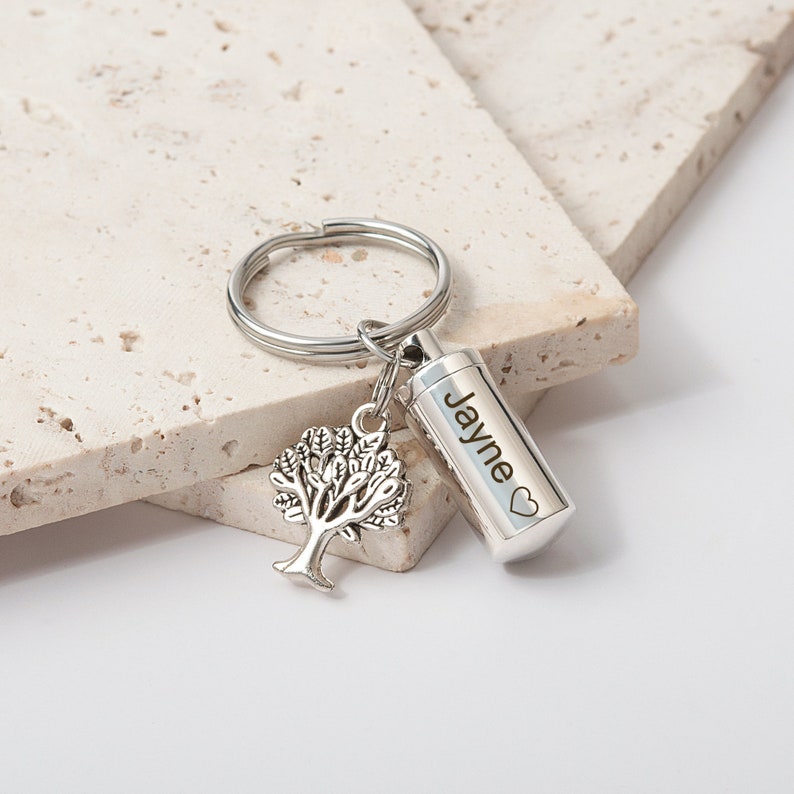 May include: Silver keychain with a small tree charm and a cylindrical cremation urn engraved with the name "Jayne" and a heart.