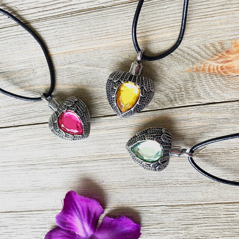 May include: Three silver heart-shaped pendants with angel wings and different colored glass stones. The pendants are on black cords and are displayed on a wooden surface.