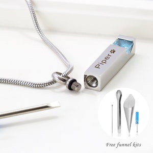 May include: Silver necklace with a rectangular pendant that has a blue stone and the inscription "Piper" with a paw print. The pendant is open and a small screwdriver and funnel are included for filling.
