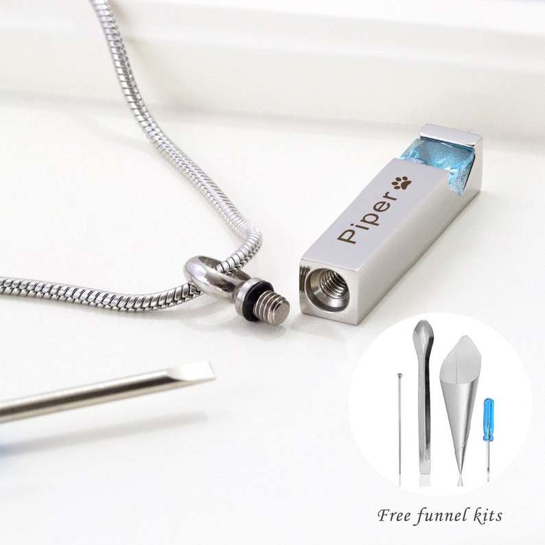 May include: Silver necklace with a rectangular pendant that has a blue stone and the inscription "Piper" with a paw print. The pendant is open and a small screwdriver and funnel are included for filling.