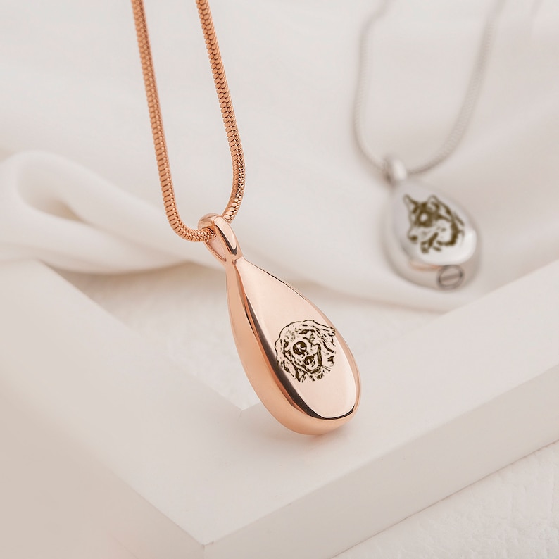 May include: Rose gold teardrop-shaped cremation necklace with a personalised engraved image of a dog. The necklace is on a white surface.