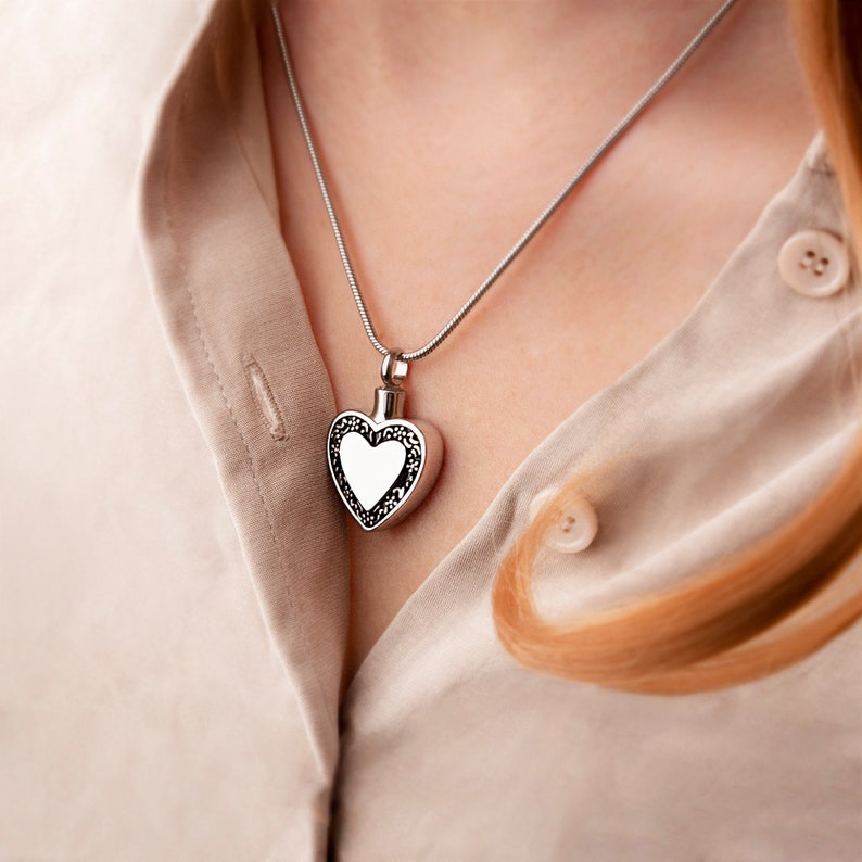 May include: A silver heart-shaped pendant necklace with a smooth, white surface inside the heart. The necklace is worn by a person with long, light brown hair.