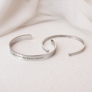Op de afbeelding: Twee zilveren armbandjes met gegraveerde berichten. Een armbandje heeft de tekst "You Are My Entire Universe" en het andere "4.22.2018 S♡L".