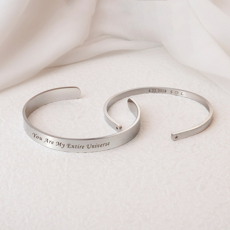 Op de afbeelding: Twee zilveren armbandjes met gegraveerde berichten. Een armbandje heeft de tekst "You Are My Entire Universe" en het andere "4.22.2018 S♡L".