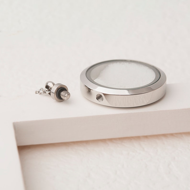 Pode incluir: Um medalh&atilde;o de prata com um inserto de vidro transparente e uma tampa de rosca. O medalh&atilde;o est&aacute; separado de uma corrente de prata e um conector de rosca.