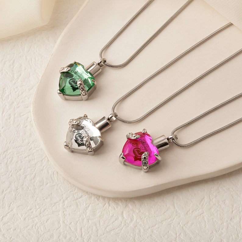 May include: Three silver heart-shaped necklaces with crystal pendants. The pendants are filled with a clear liquid and have the words "Always in My Heart" inscribed on them. The necklaces are on a white surface.