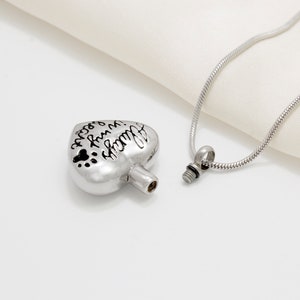 May include: Silver heart-shaped cremation necklace with a screw-on lid. The heart has the inscription "Always in my heart" and paw prints. The necklace is on a silver chain.