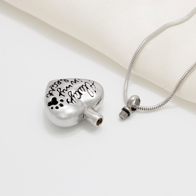 May include: Silver heart-shaped cremation necklace with a screw-on lid. The heart has the inscription "Always in my heart" and paw prints. The necklace is on a silver chain.