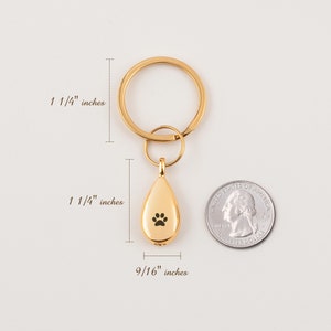 Może przedstawiać: Złoty brelok do kluczy z wisiorkiem w kształcie łzy z odciskiem łapy. Wisiorek ma 3,2 cm wysokości i 1,4 cm szerokości. Brelok ma średnicę 3,2 cm.