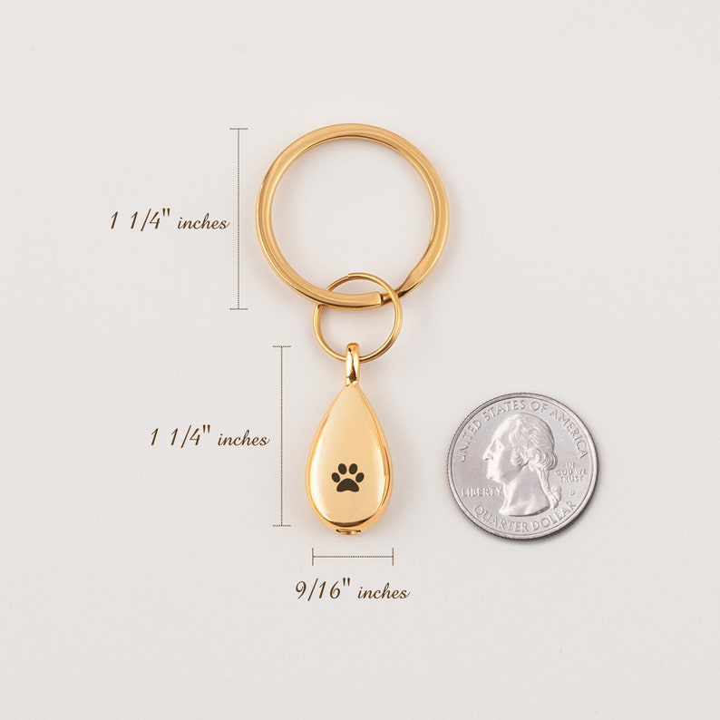 Może przedstawiać: Złoty brelok do kluczy z wisiorkiem w kształcie łzy z odciskiem łapy. Wisiorek ma 3,2 cm wysokości i 1,4 cm szerokości. Brelok ma średnicę 3,2 cm.