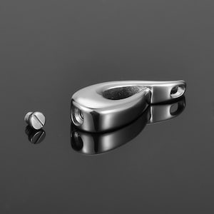 May include: A silver teardrop-shaped cremation pendant with a screw-on lid. The pendant is open and the screw is lying next to it.