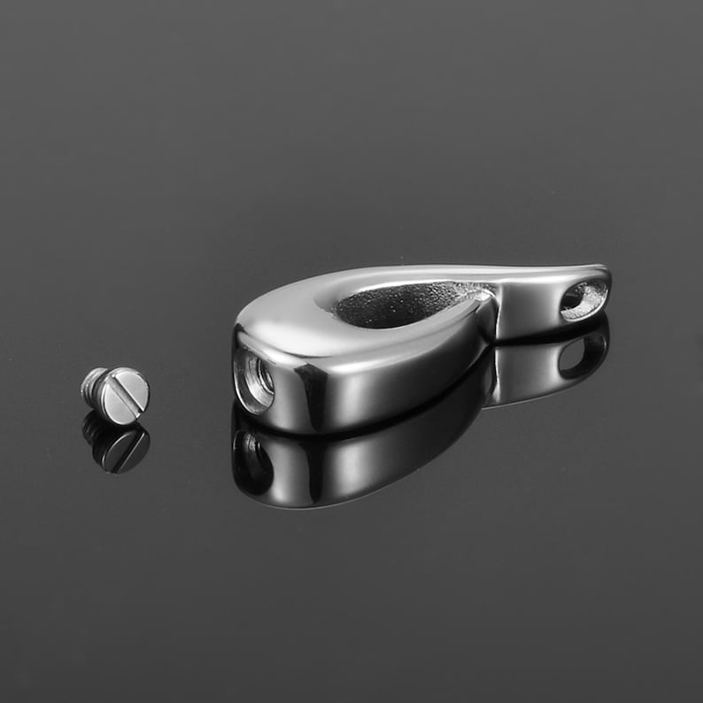May include: A silver teardrop-shaped cremation pendant with a screw-on lid. The pendant is open and the screw is lying next to it.