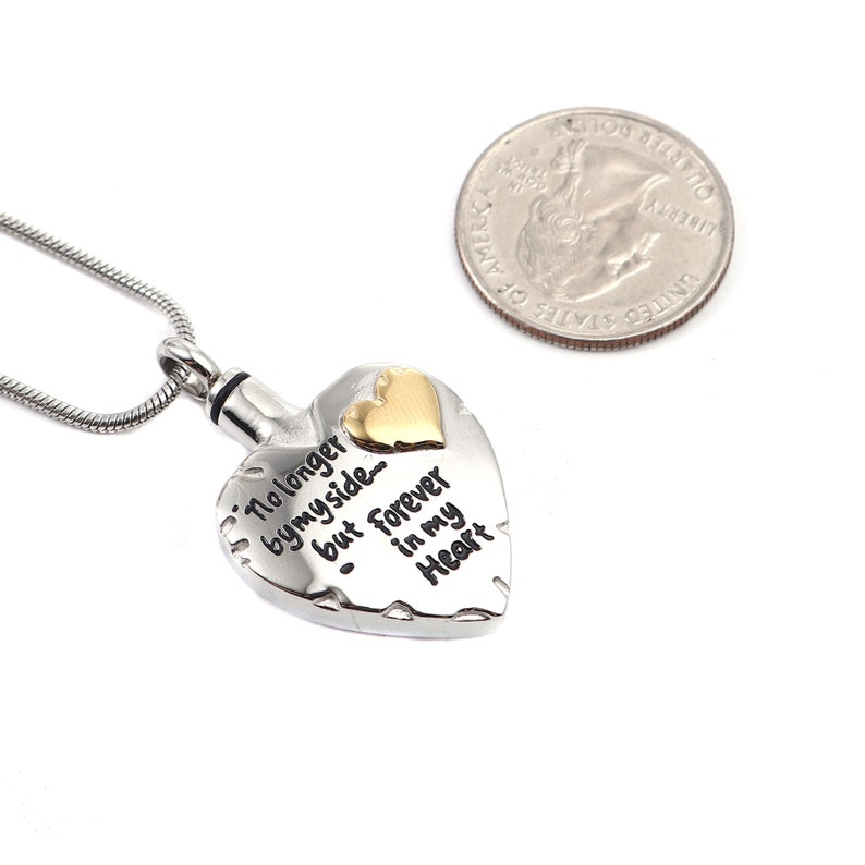 May include: Silver heart-shaped cremation necklace with a gold heart accent. The pendant has the inscription "No longer by my side, but forever in my heart".
