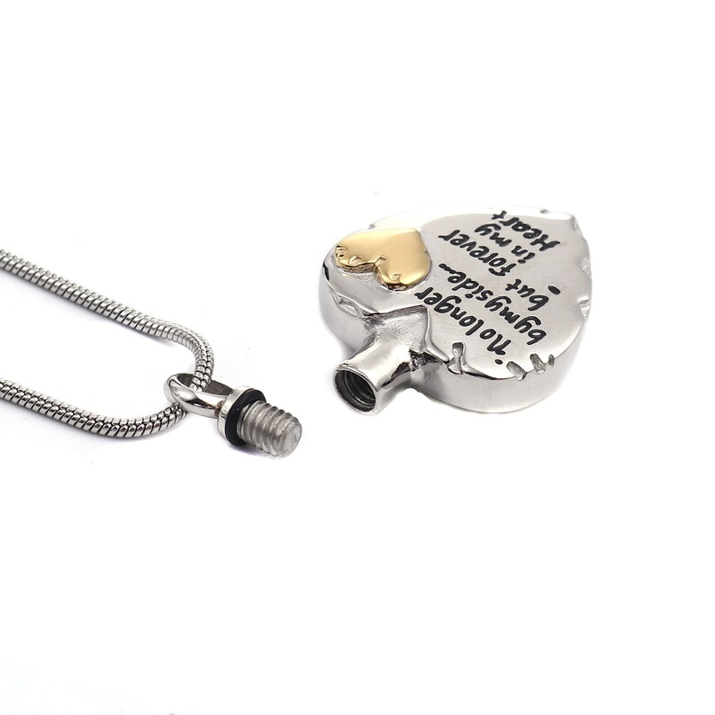 May include: Silver heart-shaped cremation necklace with a gold heart charm. The necklace has a threaded screw closure and a chain. The pendant has the inscription "No longer beside me, but forever in my heart."