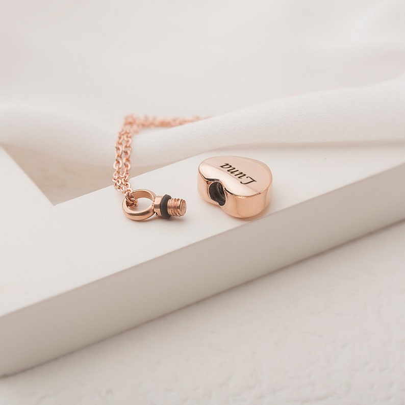 May include: Rose gold heart-shaped cremation necklace with a screw-on lid. The necklace is engraved with the name "Luna".