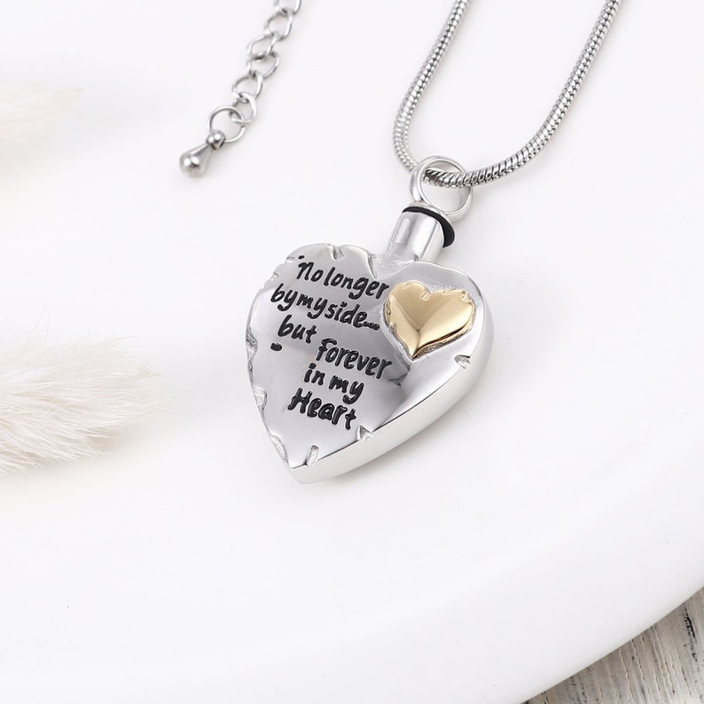 May include: Silver heart-shaped cremation necklace with a gold heart charm. The necklace has a chain and a small pendant. The heart has the inscription "No longer by my side... but forever in my heart".