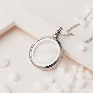 May include: A silver locket necklace with a round, clear glass insert. The necklace is lying on a white surface with small white stones.