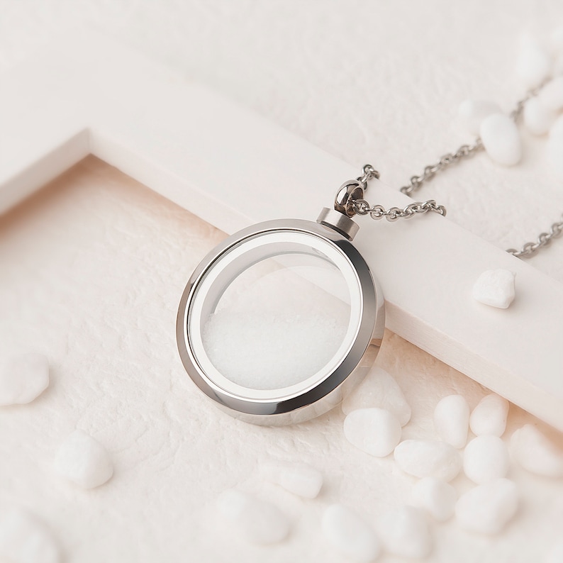 Pode incluir: Um colar de prata com um medalh&atilde;o redondo de vidro transparente. O colar est&aacute; sobre uma superf&iacute;cie branca com pequenas pedras brancas.