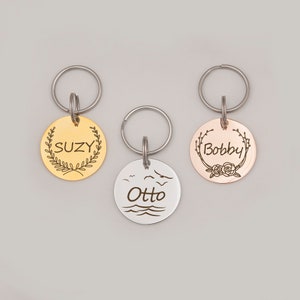 Op de afbeelding: Drie hondenpenningen in goud, zilver en ros&eacute; goud met de gegraveerde namen "Suzy", "Otto" en "Bobby". Elke penning heeft een ander ontwerp, waaronder een krans, een zeelandschap en een bloemenmotief.