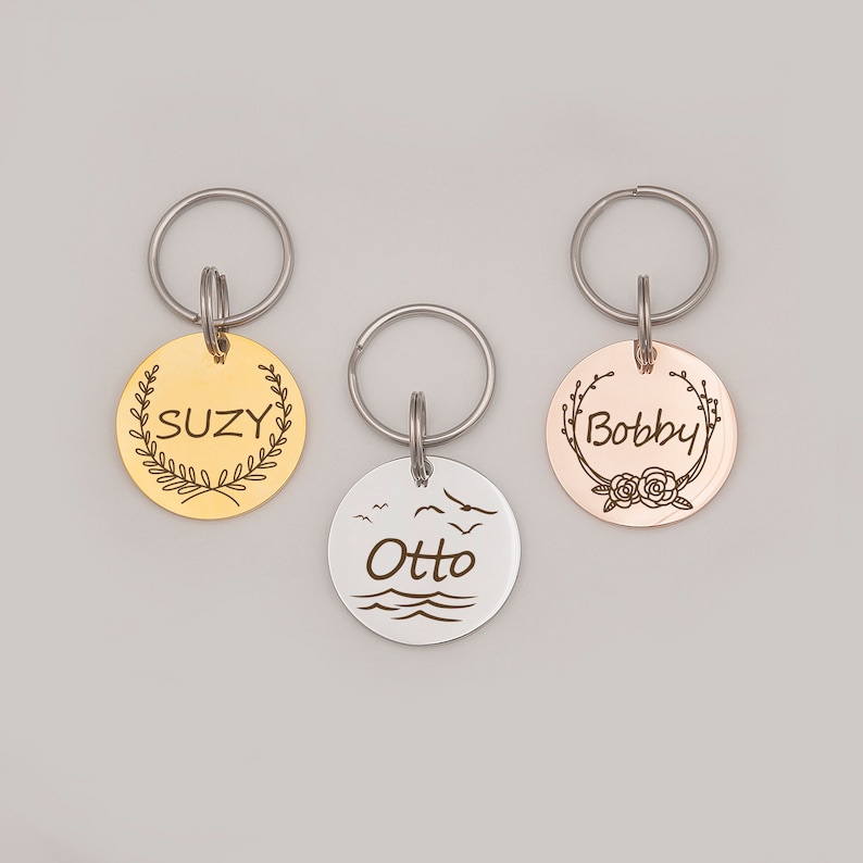 Op de afbeelding: Drie hondenpenningen in goud, zilver en ros&eacute; goud met de gegraveerde namen "Suzy", "Otto" en "Bobby". Elke penning heeft een ander ontwerp, waaronder een krans, een zeelandschap en een bloemenmotief.