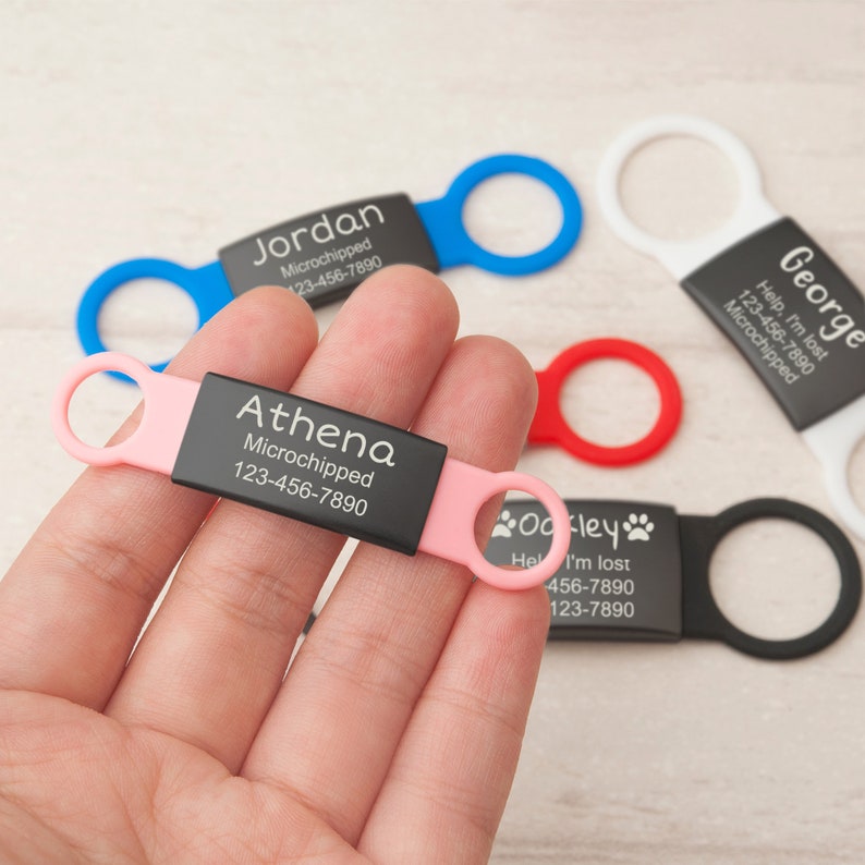 May include: A rectangular black pet ID tag with a pink silicone loop on each side. The tag is engraved with the name Athena, the word Microchipped, and the phone number 123-456-7890. Other tags in blue, white, red, and black are visible nearby.