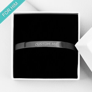 Op de afbeelding: Een zwarte metalen armband met de woorden "CUSTOM ME" gegraveerd erop. De armband zit in een witte doos met een zwarte binnenkant.