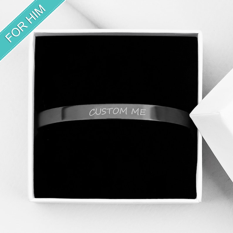 Op de afbeelding: Een zwarte metalen armband met de woorden "CUSTOM ME" gegraveerd erop. De armband zit in een witte doos met een zwarte binnenkant.