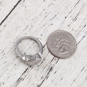 May include: A silver ring with a heart-shaped design and a screw on the side. The ring is next to a US quarter for size comparison.