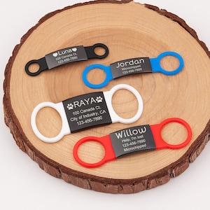 May include: Four pet ID tags with black metal centers and colored silicone bands in black, blue, white, and red. Each tag features engraved text including names like Luna, Jordan, Raya, and Willow, along with addresses and phone numbers.